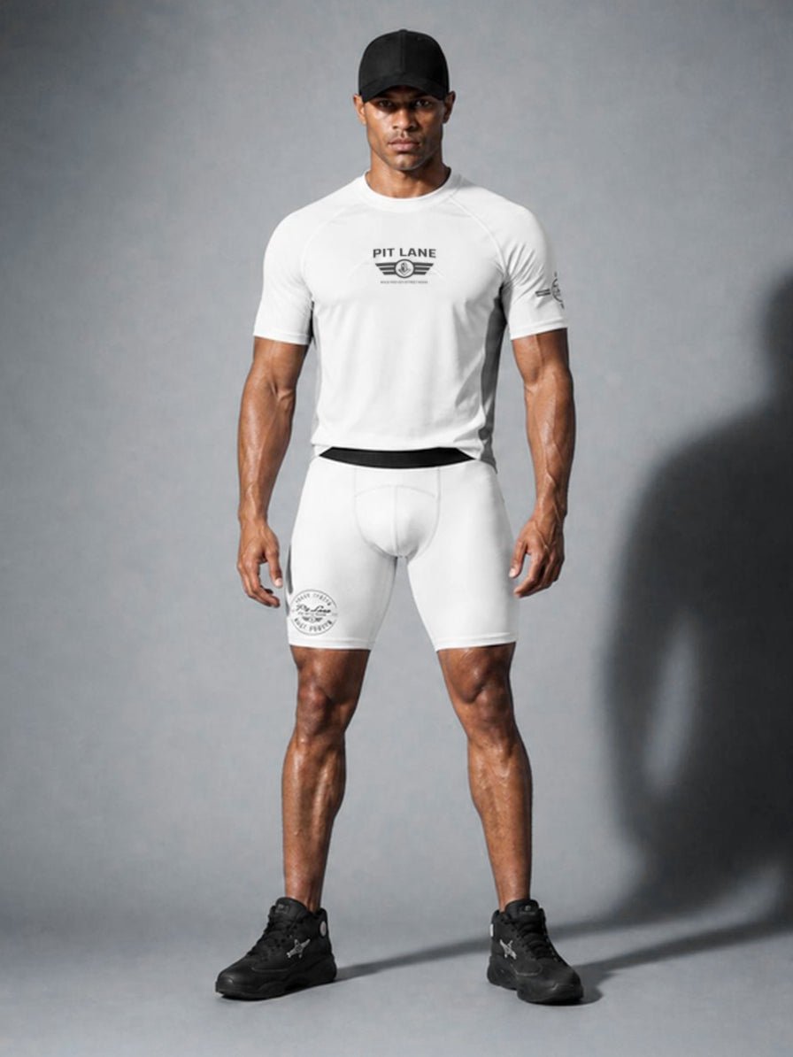 A muscular man modeling a white PIT LANE CLOTHING short-sleeve athletic t-shirt and matching white compression shorts with black branding, paired with a black cap and black sneakers, against a gray studio background.
