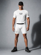 A muscular man modeling a white PIT LANE CLOTHING short-sleeve athletic t-shirt and matching white compression shorts with black branding, paired with a black cap and black sneakers, against a gray studio background.