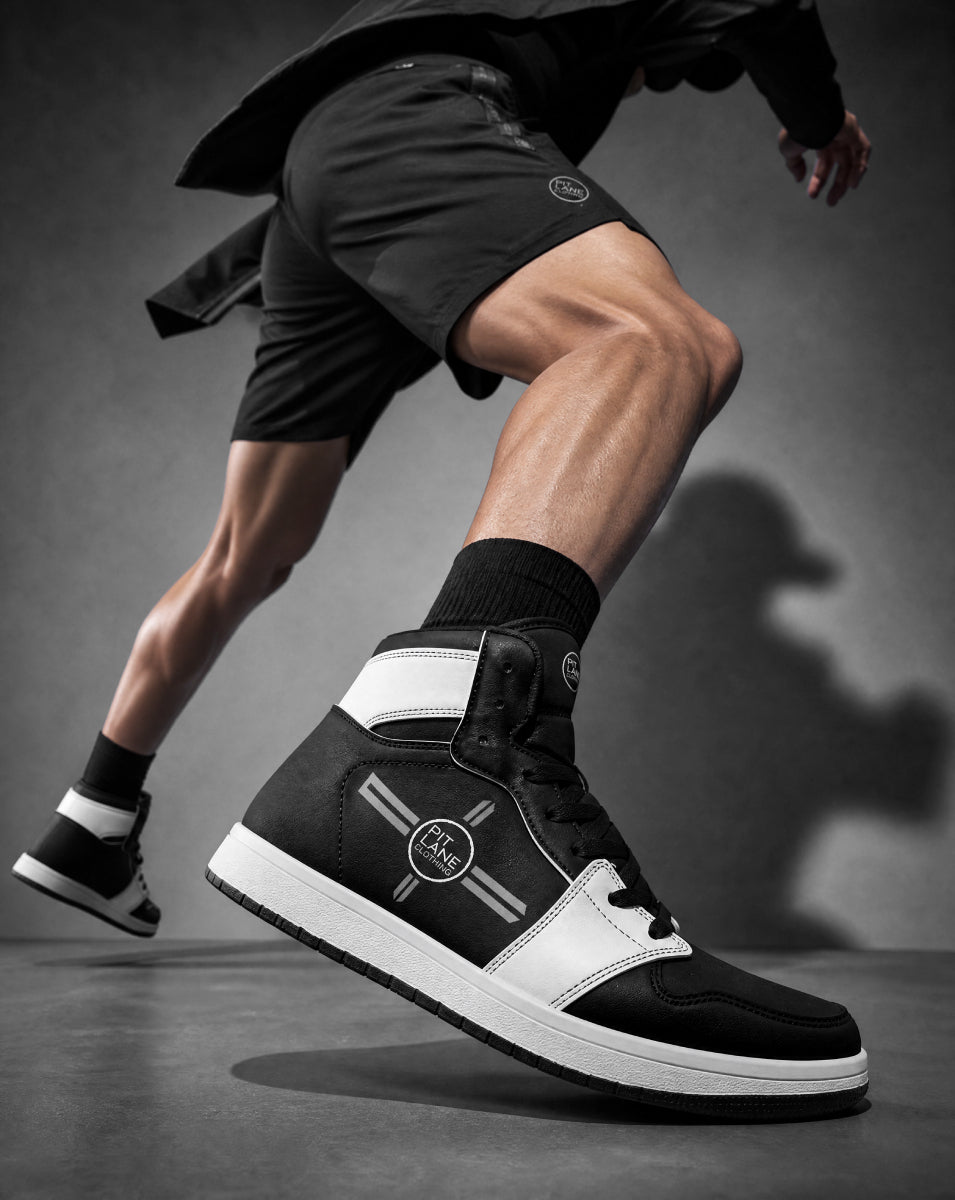 A PIT LANE CLOTHING high-top sneaker in black and white, worn by a model in a dynamic running pose against a dark blue-gray studio background.