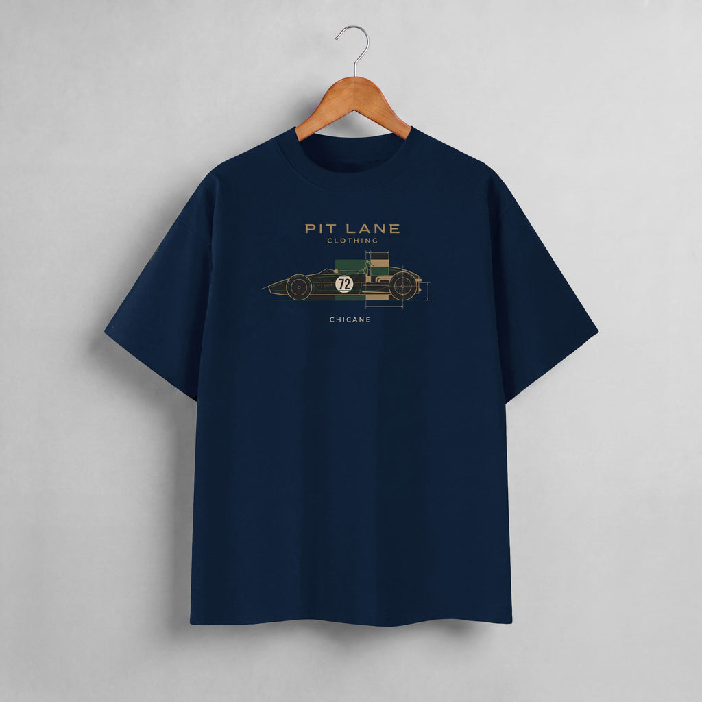 A navy blue crewneck t-shirt hanging on a wooden hanger against a light gray wall. The shirt features a graphic of a vintage race car with dimensions and the text 'PIT LANE CLOTHING', 'CHICANE', and the number '72' in gold, green, and white.