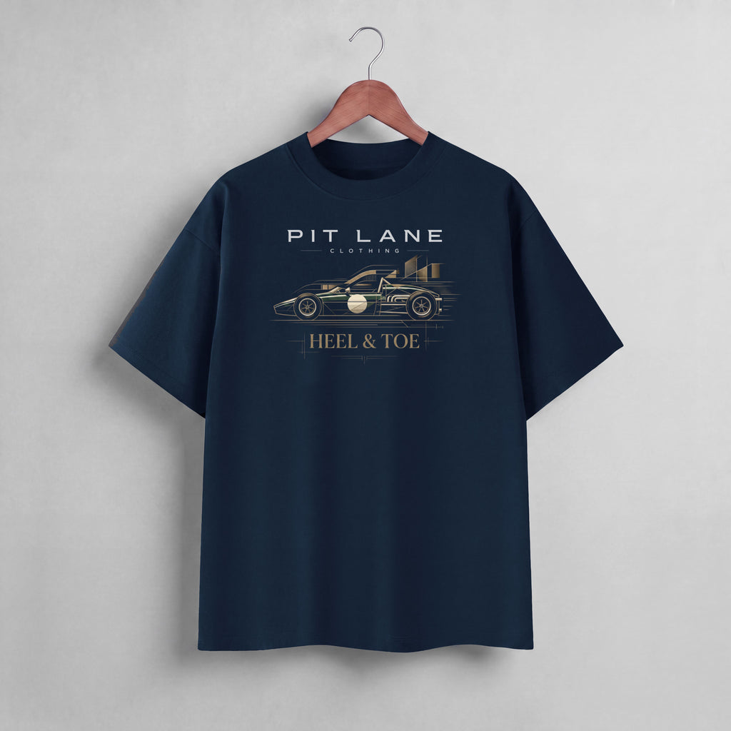 A navy blue short-sleeve t-shirt hanging on a wooden hanger against a light gray wall. The shirt features a graphic print of a race car and the text 'PIT LANE LEGENDS' and 'HEEL & TOE' in white and gold.