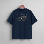 A navy blue short-sleeve t-shirt hanging on a wooden hanger against a light gray wall. The shirt features a graphic print of a race car and the text 'PIT LANE LEGENDS' and 'HEEL & TOE' in white and gold.