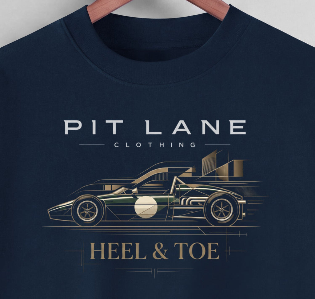 A close-up of a navy blue crewneck t-shirt hanging on a wooden hanger, featuring a graphic print of a race car and the text 'PIT LANE CLOTHING' and 'HEEL & TOE' in white and gold.