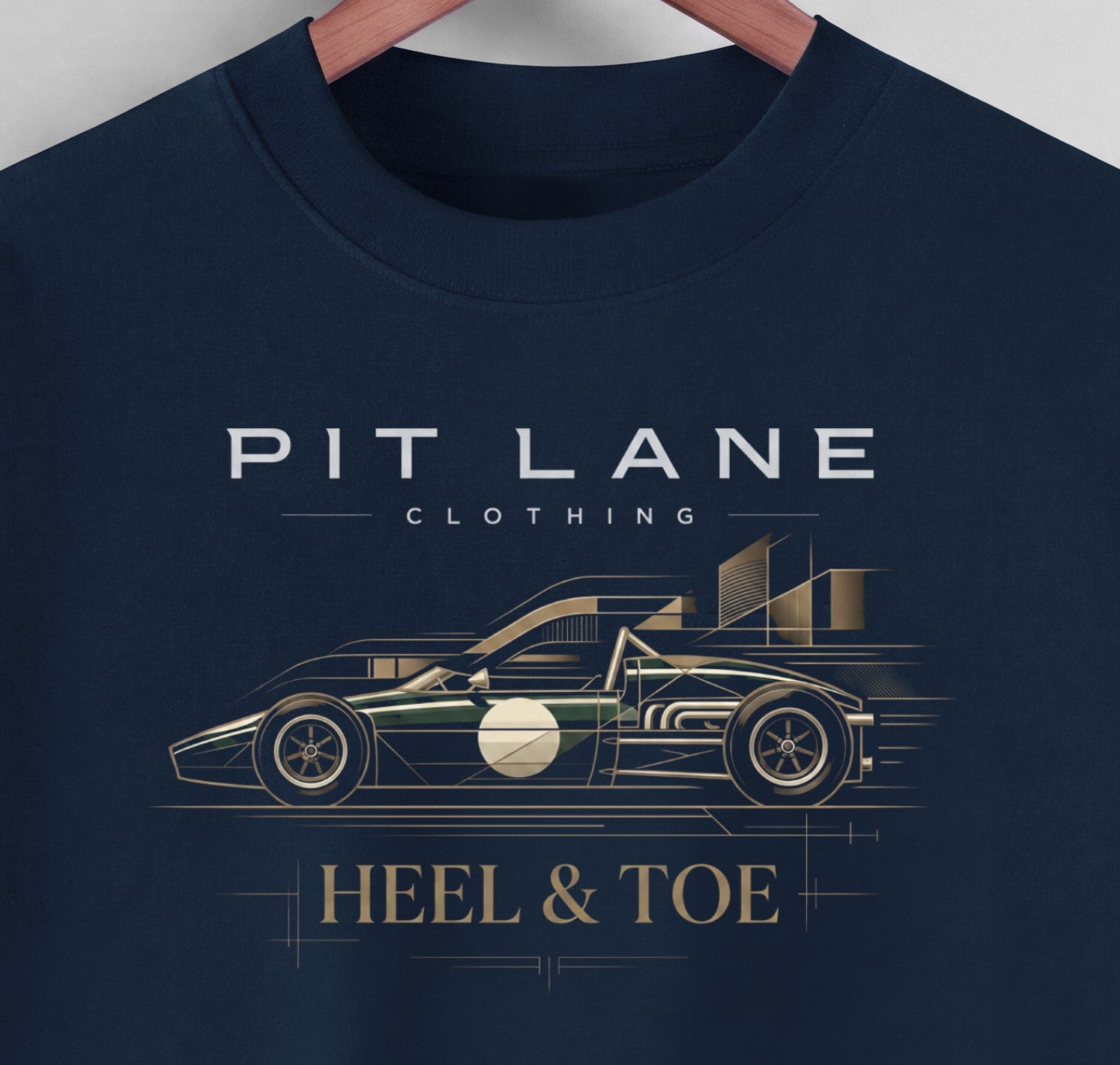 A close-up of a navy blue crewneck t-shirt hanging on a wooden hanger, featuring a graphic print of a race car and the text 'PIT LANE CLOTHING' and 'HEEL & TOE' in white and gold.