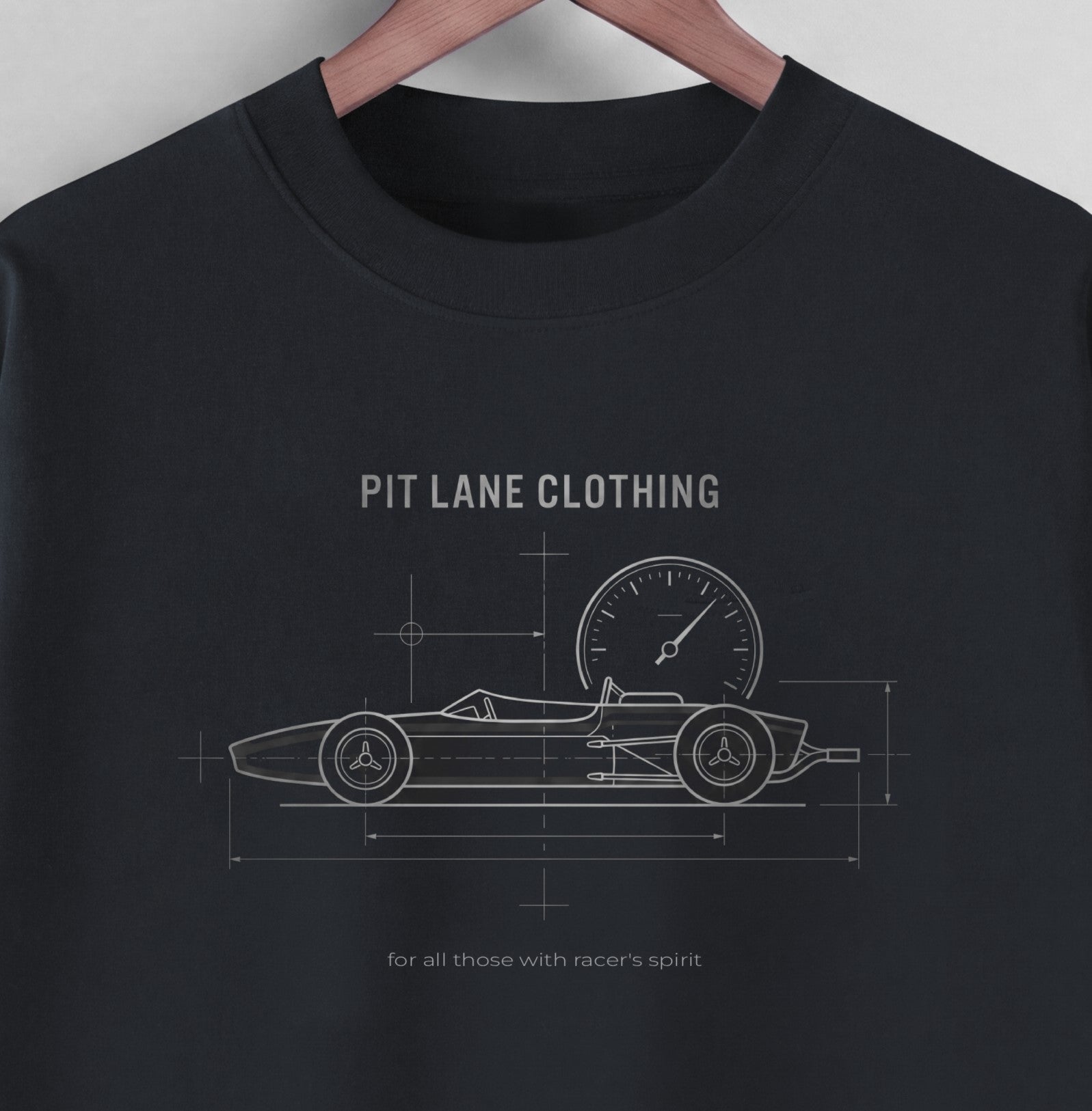 A black crewneck t-shirt hanging on a wooden hanger against a light gray wall. The shirt features a white blueprint-style graphic of a vintage race car with a speedometer, labeled 'PIT LANE CLOTHING' and 'for all those with racer's spirit'.