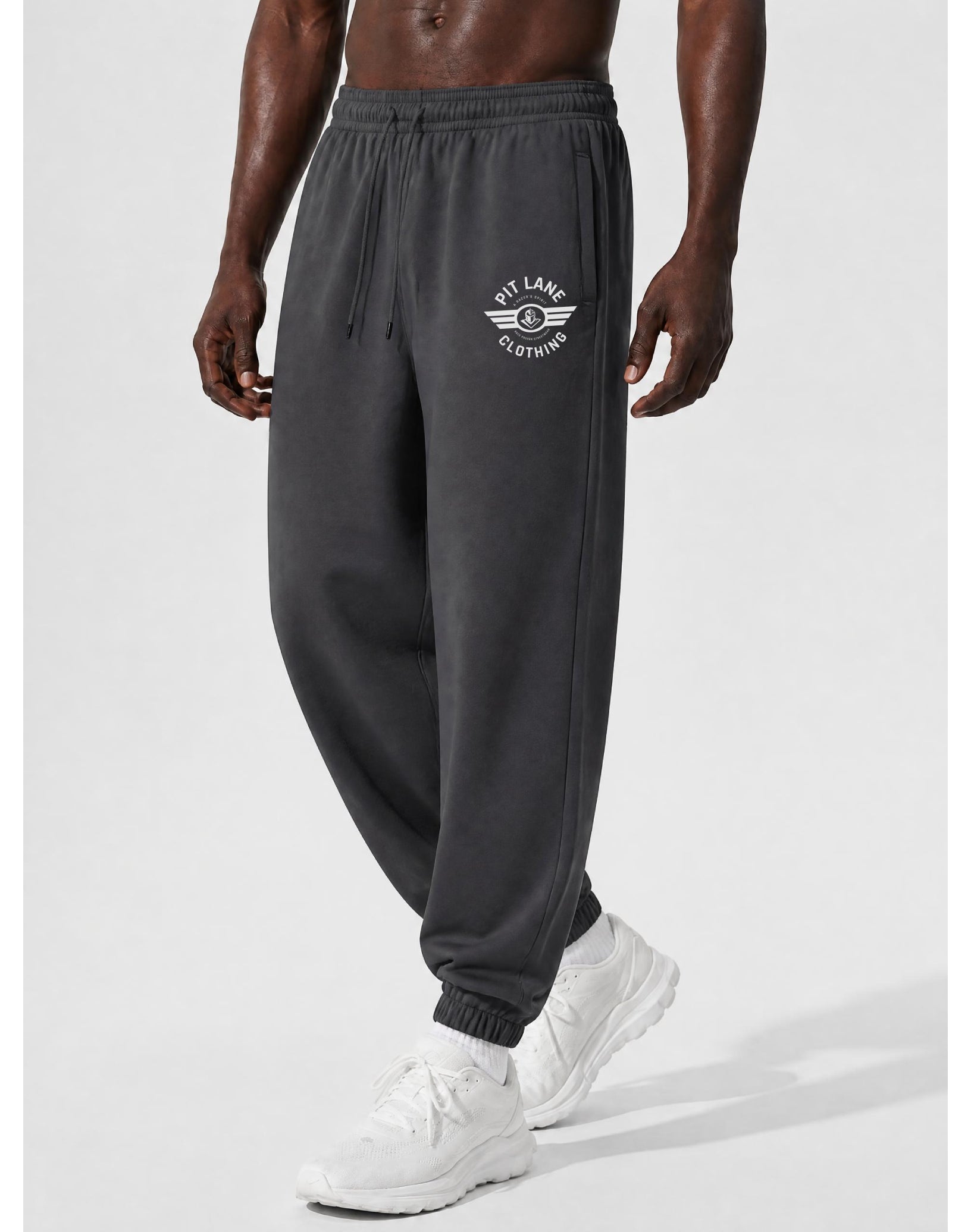 A man wearing dark gray jogger sweatpants with a white 'PIT LANE CLOTHING' winged logo on the left thigh, paired with white sneakers, shown from the waist down against a light gray studio background.