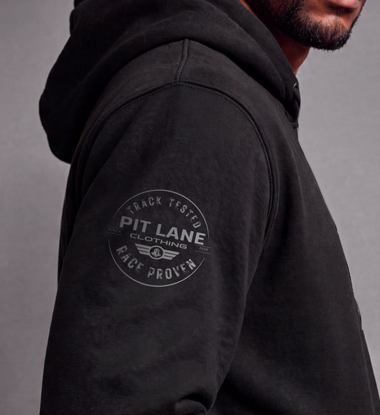 Close-up of a man wearing a black hooded sweatshirt, showing the side profile and a circular gray graphic on the upper left sleeve that reads 'PIT LANE CLOTHING' with 'TRACK TESTED' and 'RACE PROVEN'.