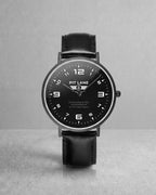 A Pit Lane Clothing analog wristwatch with a black leather strap, black dial, and silver-tone case, centered against a light gray textured background.