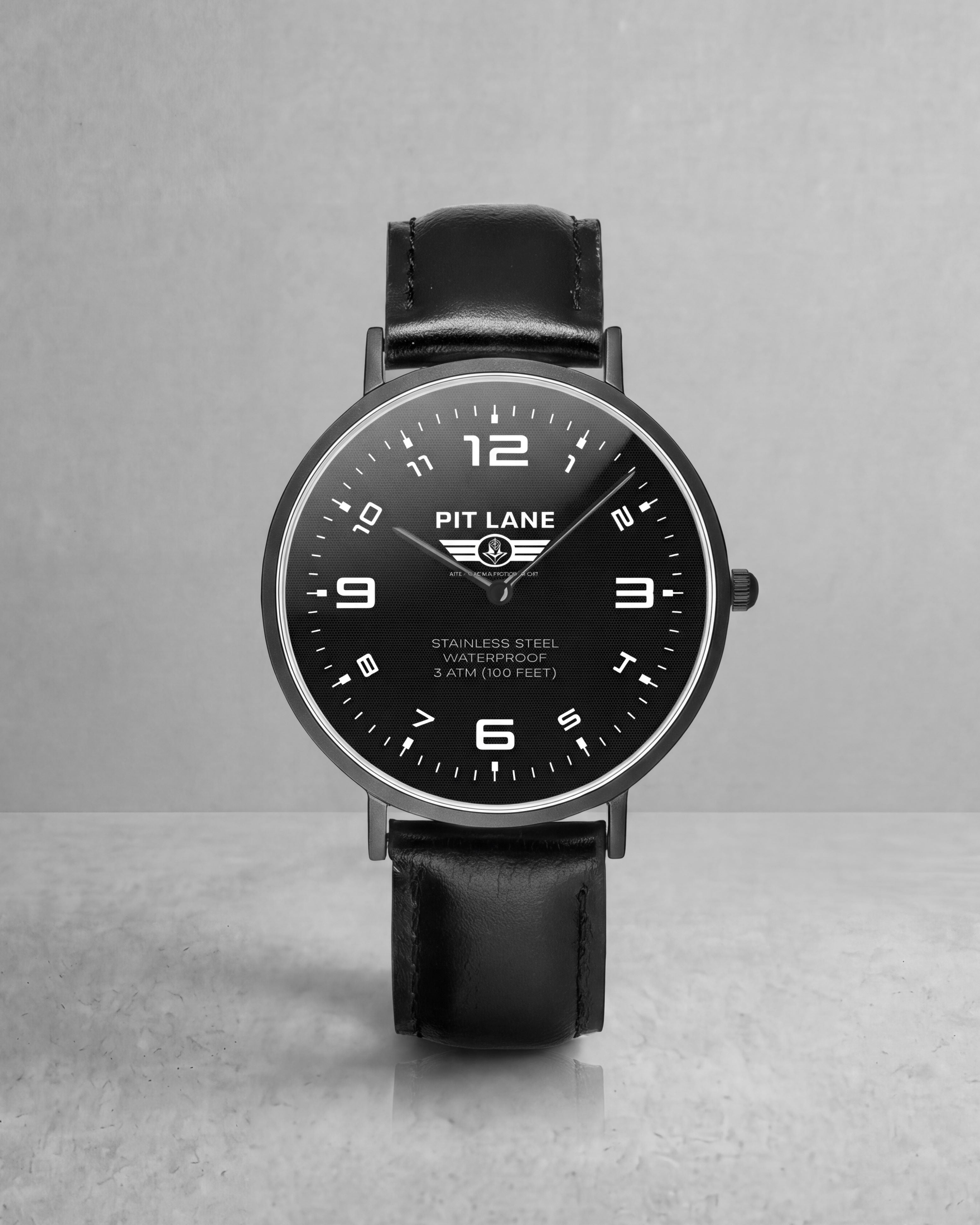 A Pit Lane Clothing analog wristwatch with a black leather strap, black dial, and silver-tone case, centered against a light gray textured background.