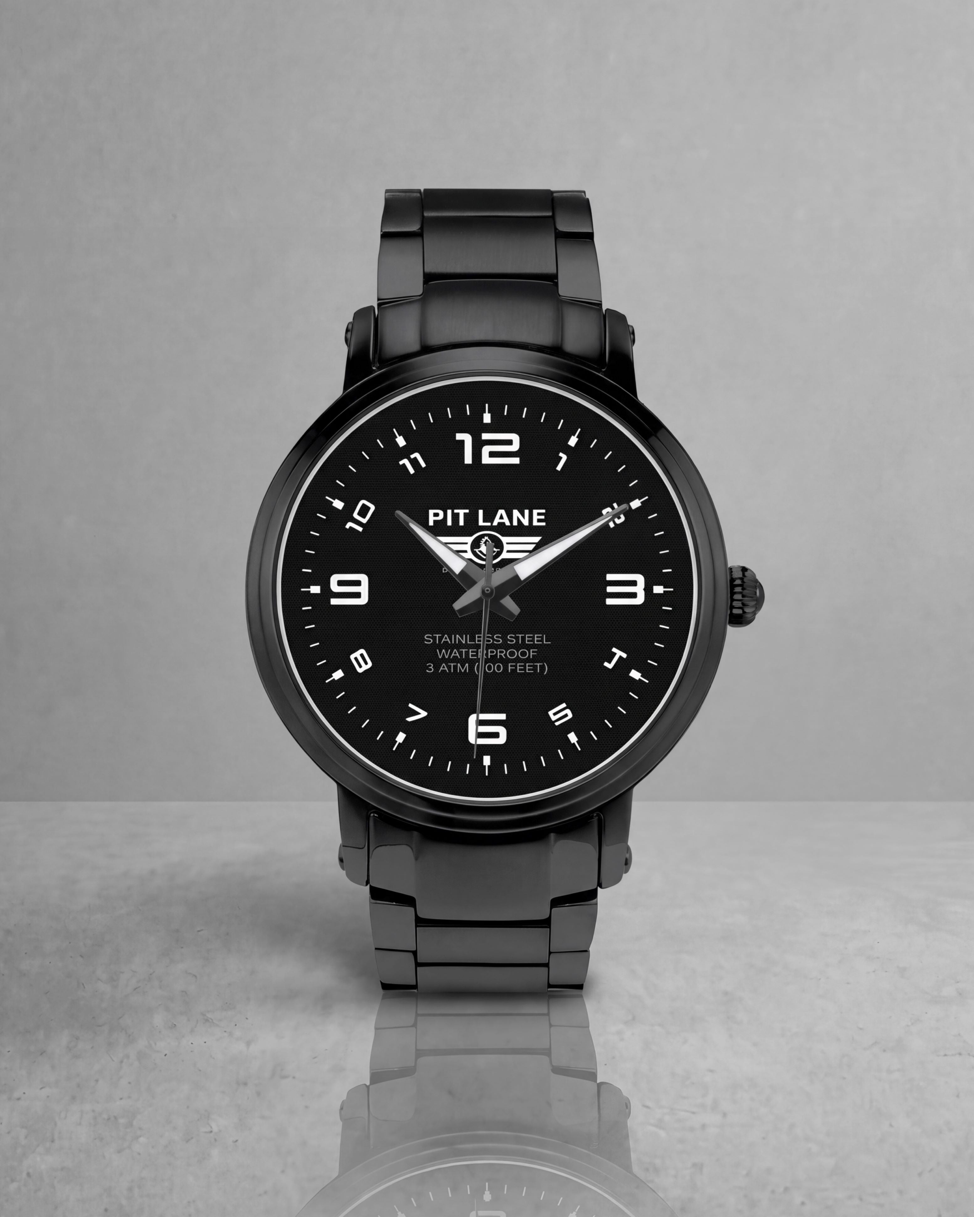 A black Pit Lane Clothing stainless steel watch with a matching black metal link bracelet, featuring a black dial with white Arabic numerals and hands, set against a neutral gray background.