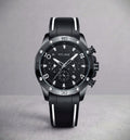 A Pitlane chronograph watch with a black dial, black bezel, and a black rubber strap with white stripes, set against a neutral gray background.
