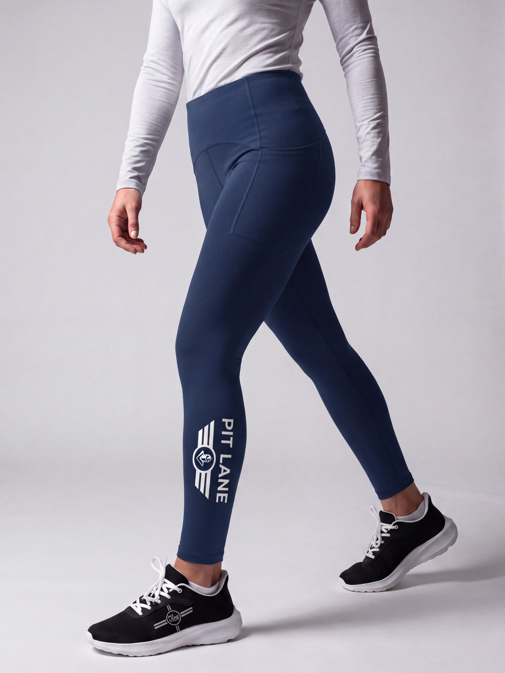 A model wearing navy blue athletic leggings with a white 'PIT LANE' logo on the lower left leg, paired with a white long-sleeve top and black running shoes, shown from the waist down against a light gray studio background.