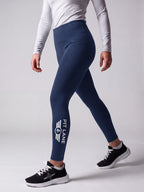 A model wearing navy blue athletic leggings with a white 'PIT LANE' logo on the lower left leg, paired with a white long-sleeve top and black running shoes, shown from the waist down against a light gray studio background.