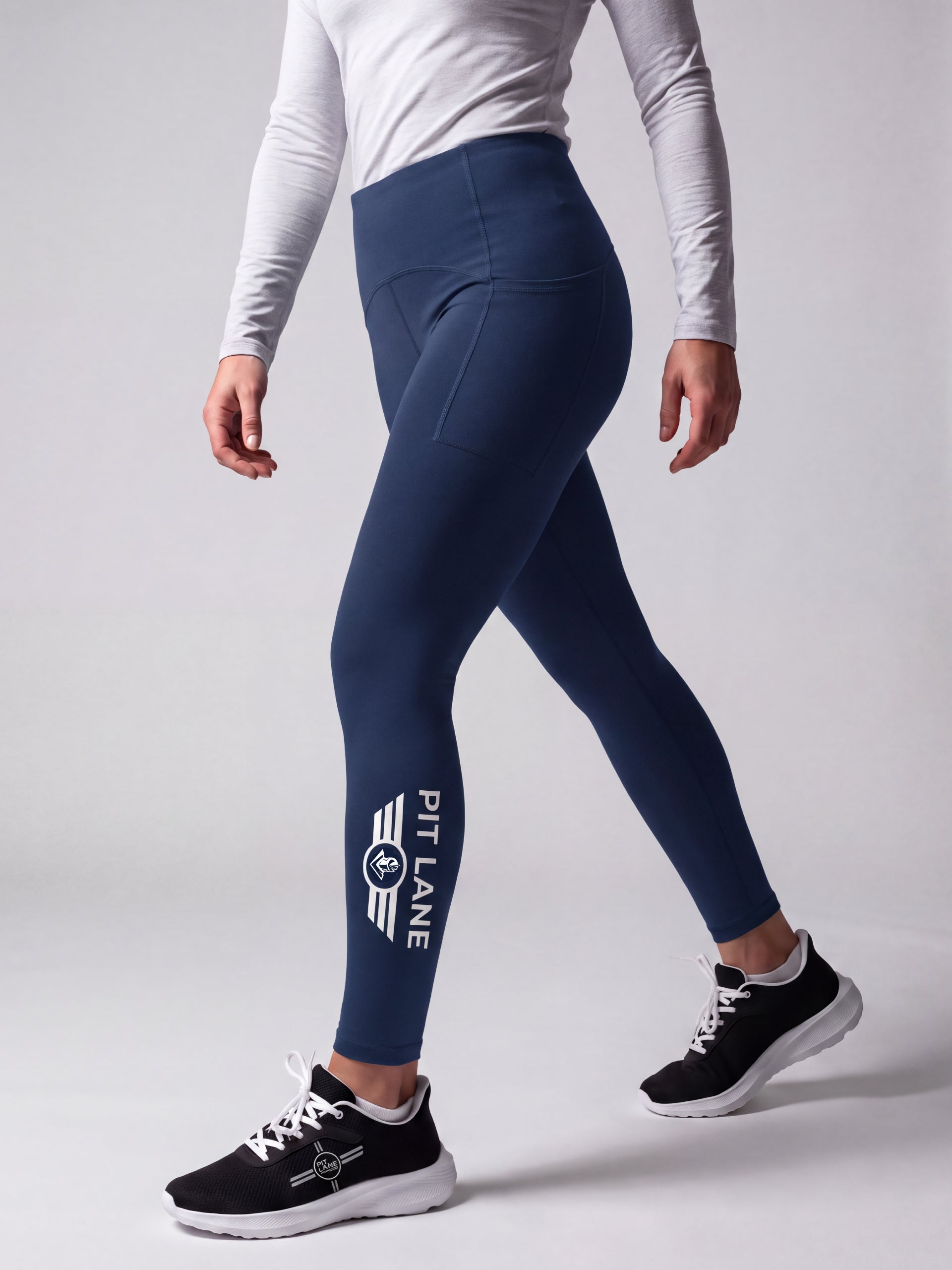 A model wearing navy blue athletic leggings with a white 'PIT LANE' logo on the lower left leg, paired with a white long-sleeve top and black running shoes, shown from the waist down against a light gray studio background.