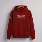 A dark red pullover hoodie hanging on a wooden hanger against a light gray wall. The hoodie features a distressed gold graphic on the chest with the text 'PIT LANE' and 'RACE DRIVER' beneath it, and a matching circular graphic on the left sleeve.