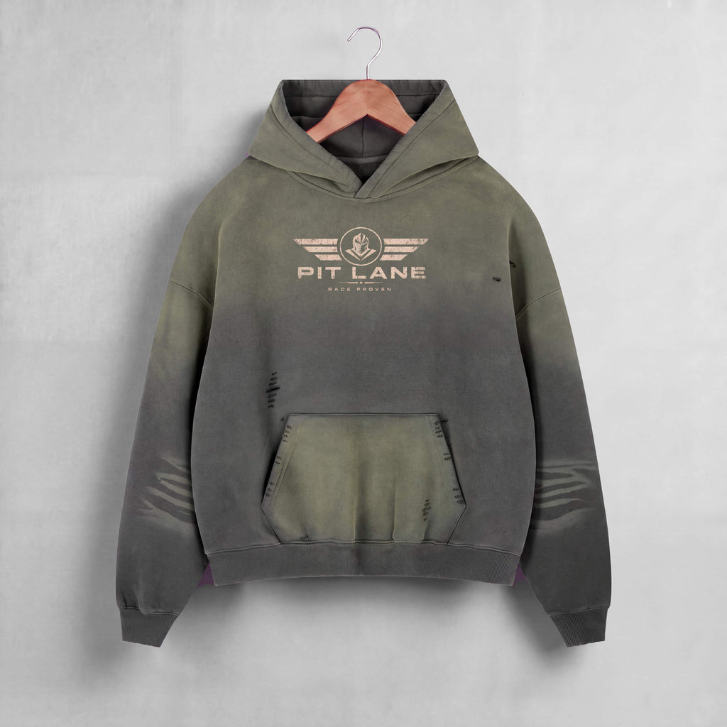A hooded sweatshirt hanging on a wooden hanger against a light gray wall. The hoodie has a distressed, faded color gradient from olive green at the top to dark charcoal gray at the bottom, with a distressed front kangaroo pocket and a 'PIT LANE' winged logo printed on the chest.