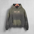 A hooded sweatshirt hanging on a wooden hanger against a light gray wall. The hoodie has a distressed, faded color gradient from olive green at the top to dark charcoal gray at the bottom, with a distressed front kangaroo pocket and a 'PIT LANE' winged logo printed on the chest.