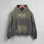 A hooded sweatshirt hanging on a wooden hanger against a light gray wall. The hoodie has a distressed, faded color gradient from olive green at the top to dark charcoal gray at the bottom, with a distressed front kangaroo pocket and a 'PIT LANE' winged logo printed on the chest.