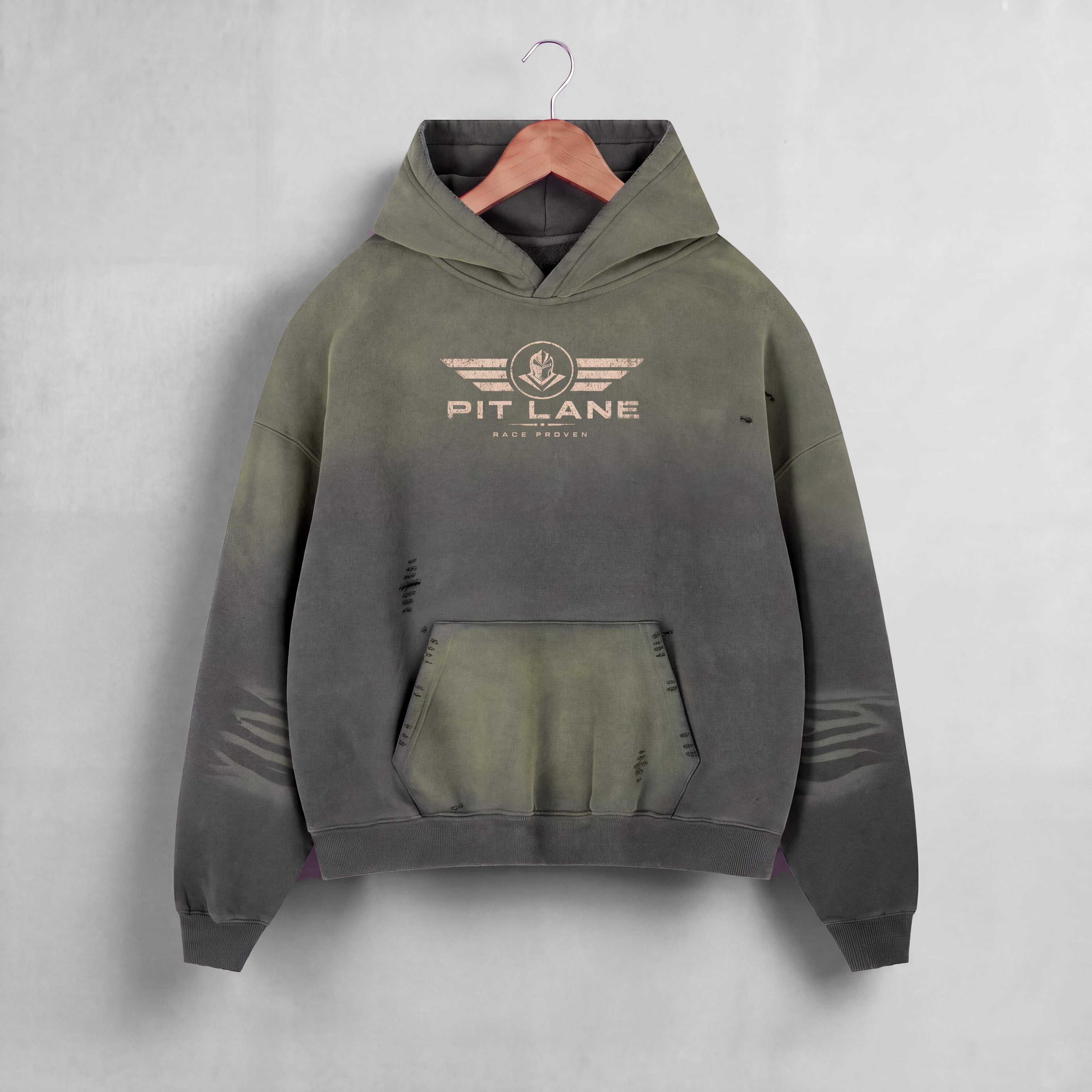 A hooded sweatshirt hanging on a wooden hanger against a light gray wall. The hoodie has a distressed, faded color gradient from olive green at the top to dark charcoal gray at the bottom, with a distressed front kangaroo pocket and a 'PIT LANE' winged logo printed on the chest.