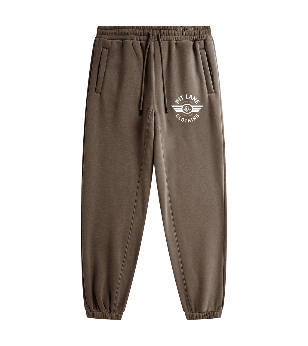 A person wearing brown jogger sweatpants with a white circular chest logo that reads 'PIT LANE CLOTHING' and white sneakers, photographed against a clean light gray studio background.