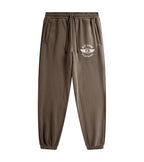 A person wearing brown jogger sweatpants with a white circular chest logo that reads 'PIT LANE CLOTHING' and white sneakers, photographed against a clean light gray studio background.