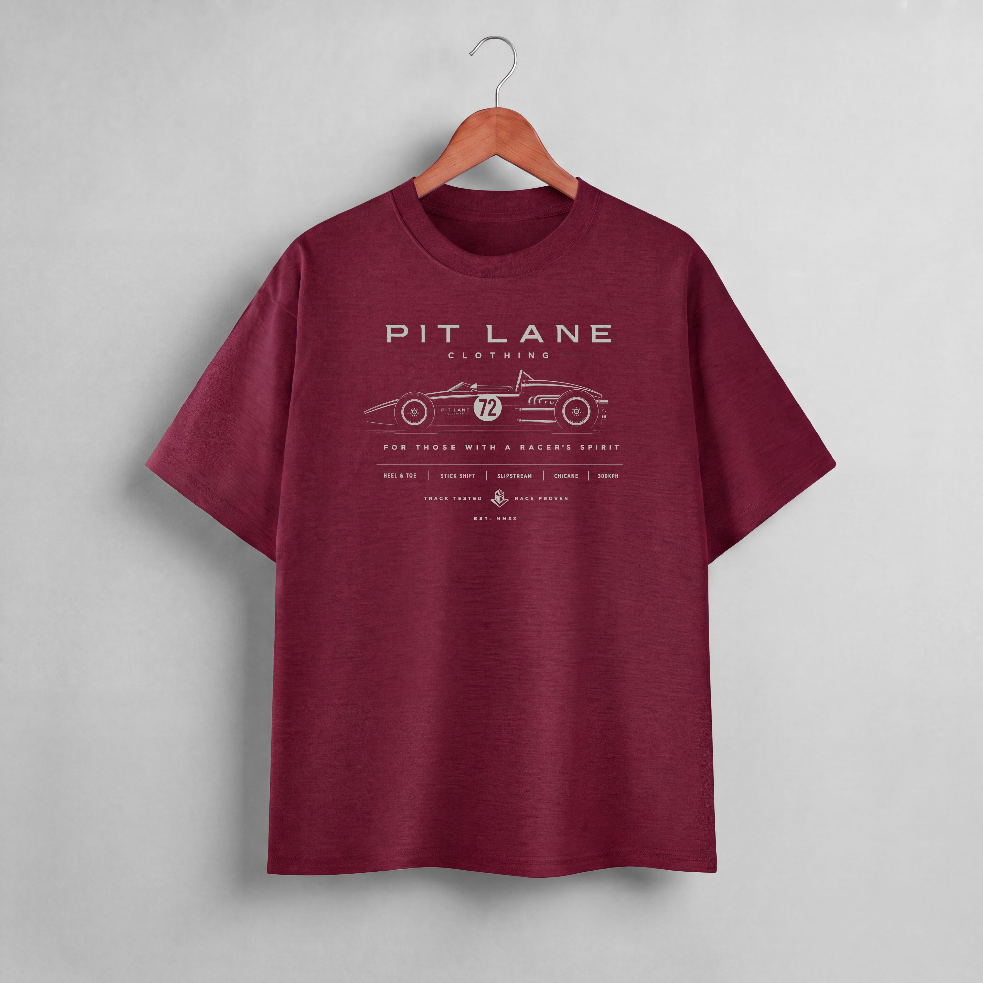 A maroon short-sleeve crewneck t-shirt hanging on a wooden hanger against a light gray wall. The shirt features a white graphic print on the chest depicting a vintage race car with the text 'PIT LANE CLOTHING' and 'WITH A RACER'S SPIRIT'.