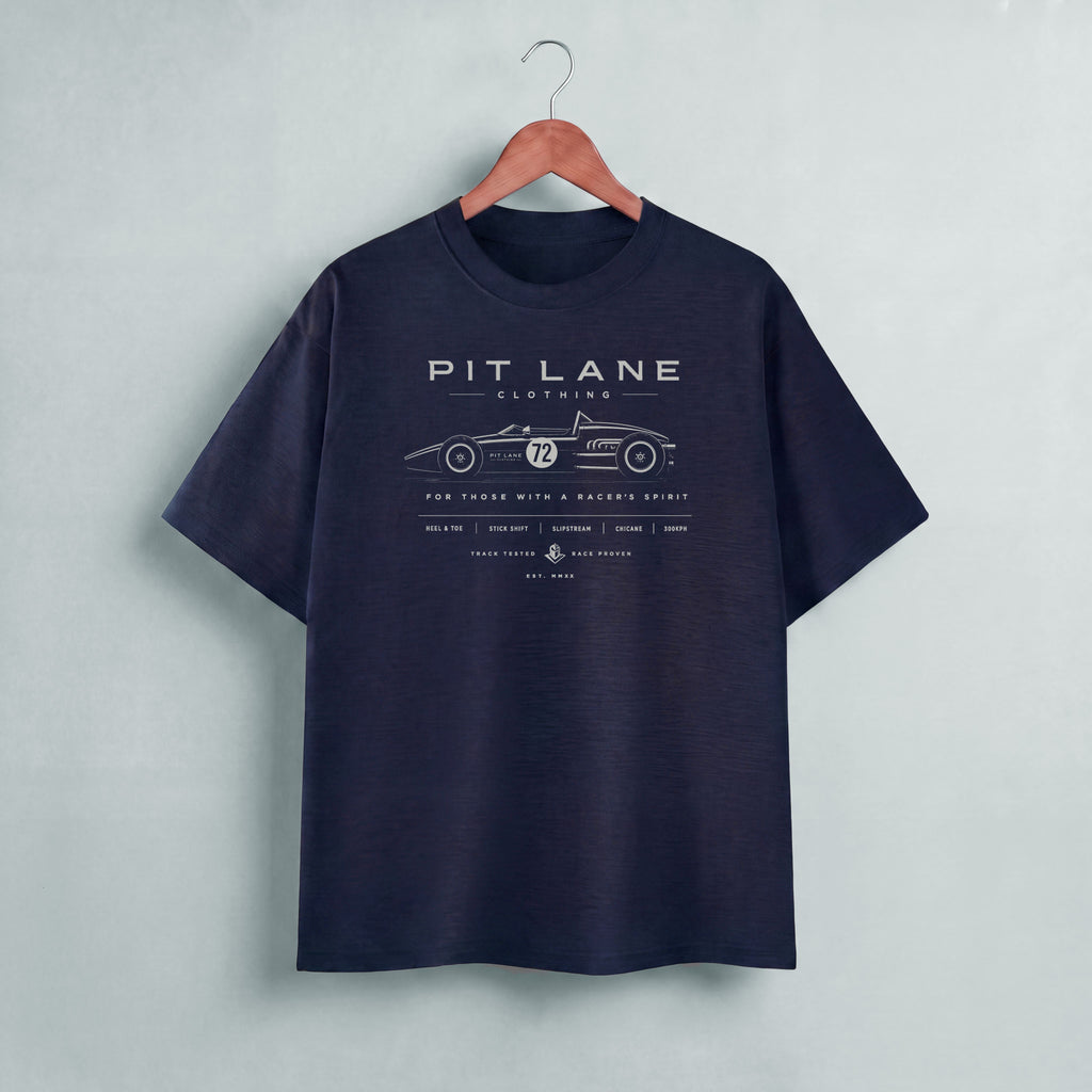 A black short-sleeve t-shirt hanging on a wooden hanger against a light gray wall. The shirt features a vintage-style race car graphic with the text 'PIT LANE CLOTHING' 