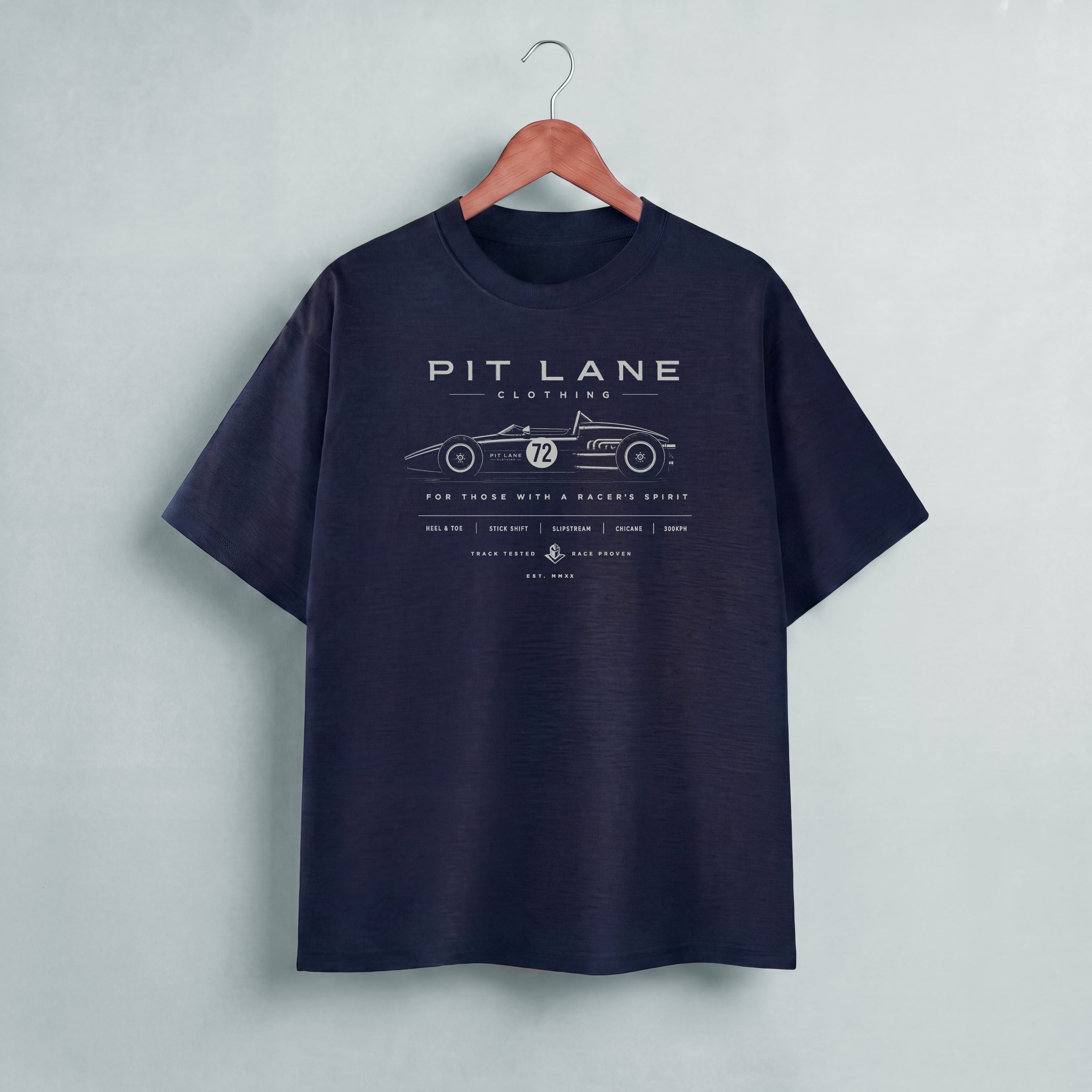 A black short-sleeve t-shirt hanging on a wooden hanger against a light gray wall. The shirt features a vintage-style race car graphic with the text 'PIT LANE CLOTHING' 