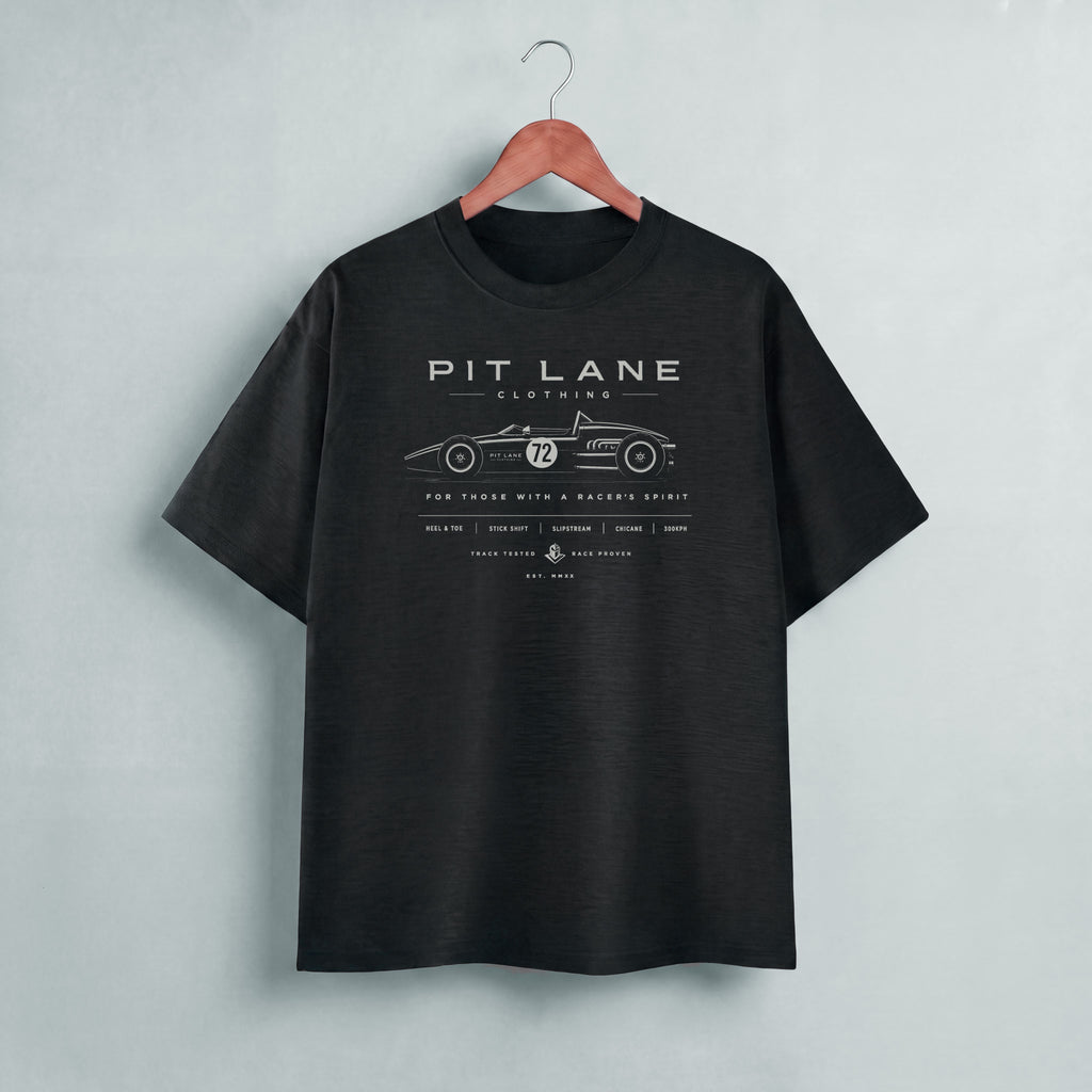 A black short-sleeve t-shirt hanging on a wooden hanger against a light gray wall. The shirt features a white graphic print of a vintage race car with the text 'PIT LANE CLOTHING' on the chest.