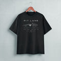 A black short-sleeve t-shirt hanging on a wooden hanger against a light gray wall. The shirt features a white graphic print of a vintage race car with the text 'PIT LANE CLOTHING' on the chest.