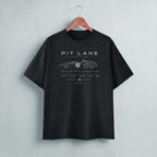 A black short-sleeve t-shirt hanging on a wooden hanger against a light gray wall. The shirt features a white graphic print of a vintage race car with the text 'PIT LANE CLOTHING' on the chest.