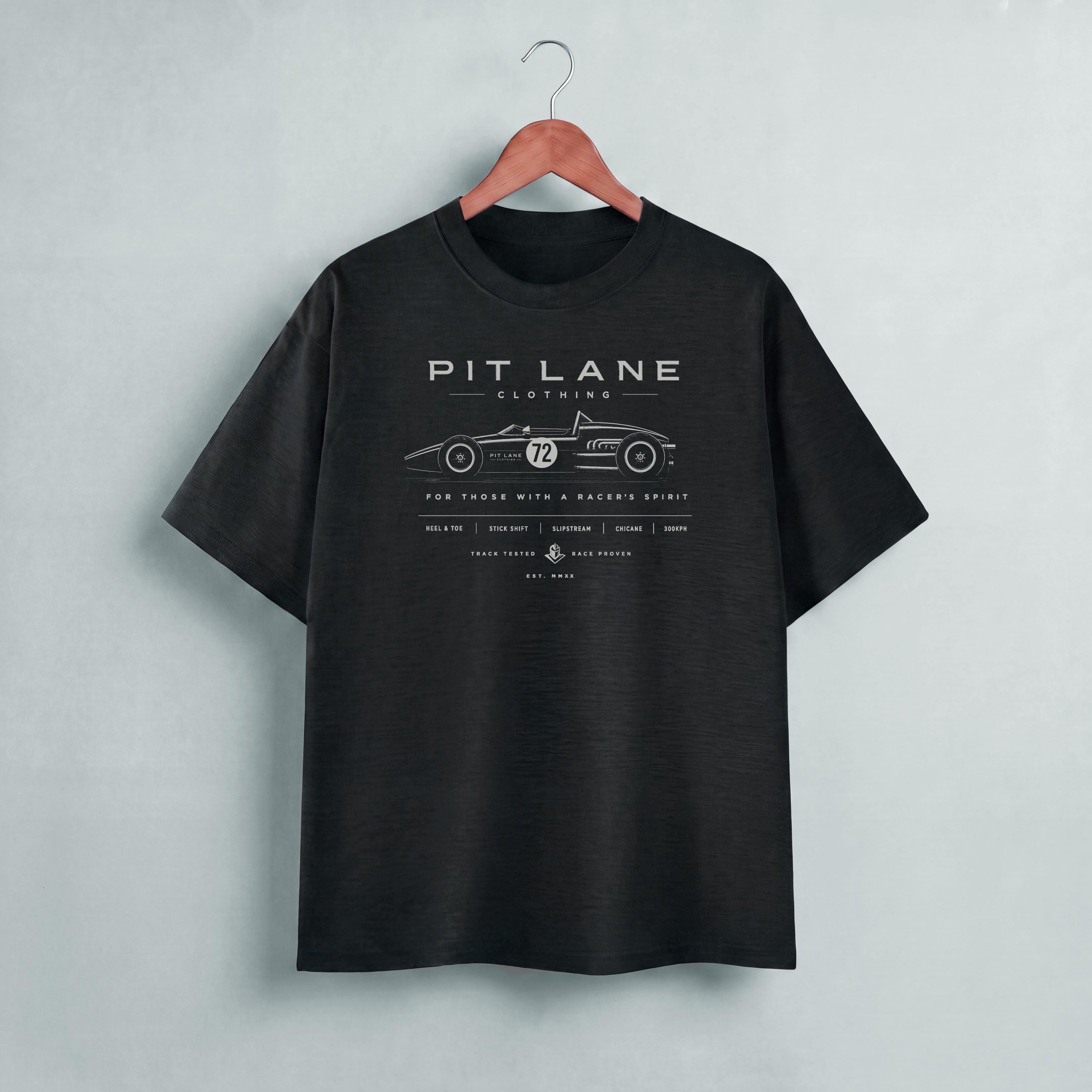 A black short-sleeve t-shirt hanging on a wooden hanger against a light gray wall. The shirt features a white graphic print of a vintage race car with the text 'PIT LANE CLOTHING' on the chest.