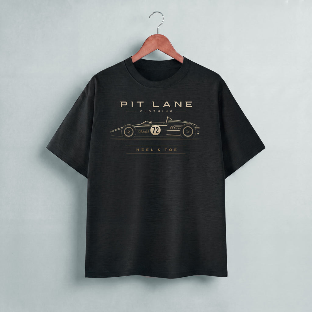 A black short-sleeve t-shirt hanging on a wooden hanger against a light gray wall. The shirt features a gold graphic print of a vintage race car with the text 'PIT LANE CLOTHING' and 'HEEL & TOE' on the chest.