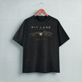 A black short-sleeve t-shirt hanging on a wooden hanger against a light gray wall. The shirt features a gold graphic print of a vintage race car with the text 'PIT LANE CLOTHING' and 'HEEL & TOE' on the chest.