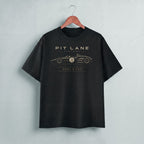 A black short-sleeve t-shirt hanging on a wooden hanger against a light gray wall. The shirt features a gold graphic print of a vintage race car with the text 'PIT LANE CLOTHING' and 'HEEL & TOE' on the chest.