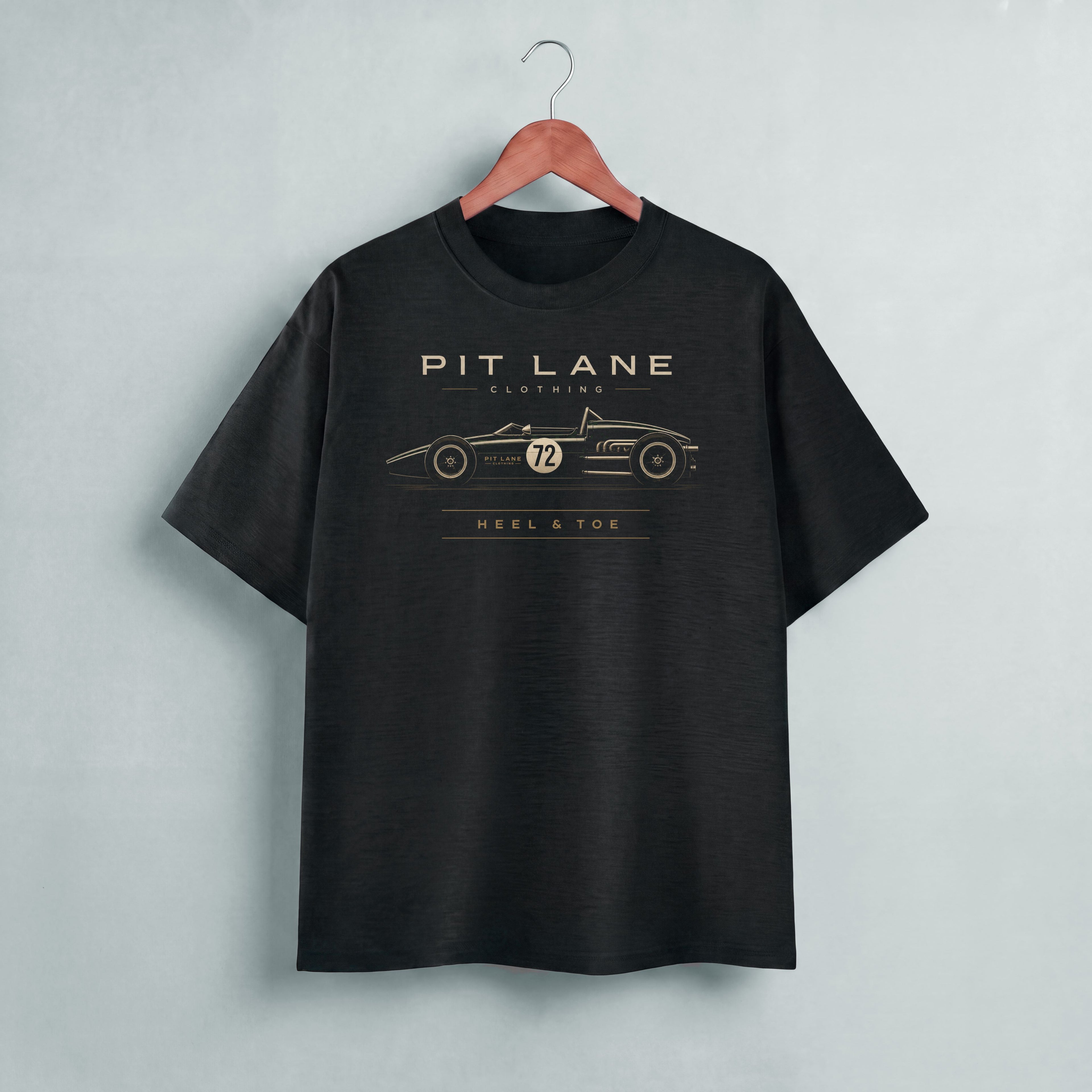A black short-sleeve t-shirt hanging on a wooden hanger against a light gray wall. The shirt features a gold graphic print of a vintage race car with the text 'PIT LANE CLOTHING' and 'HEEL & TOE' on the chest.