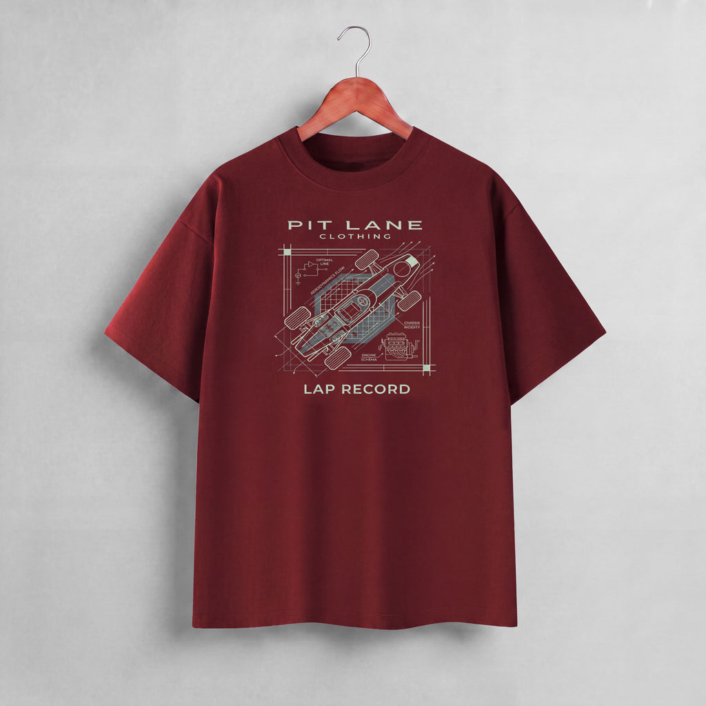 A maroon short-sleeve t-shirt hanging on a wooden hanger against a textured gray wall. The shirt features a graphic print on the front with the text 'PIT LANE CLOTHING' and 'LAP RECORD' surrounding a technical schematic of a race car.