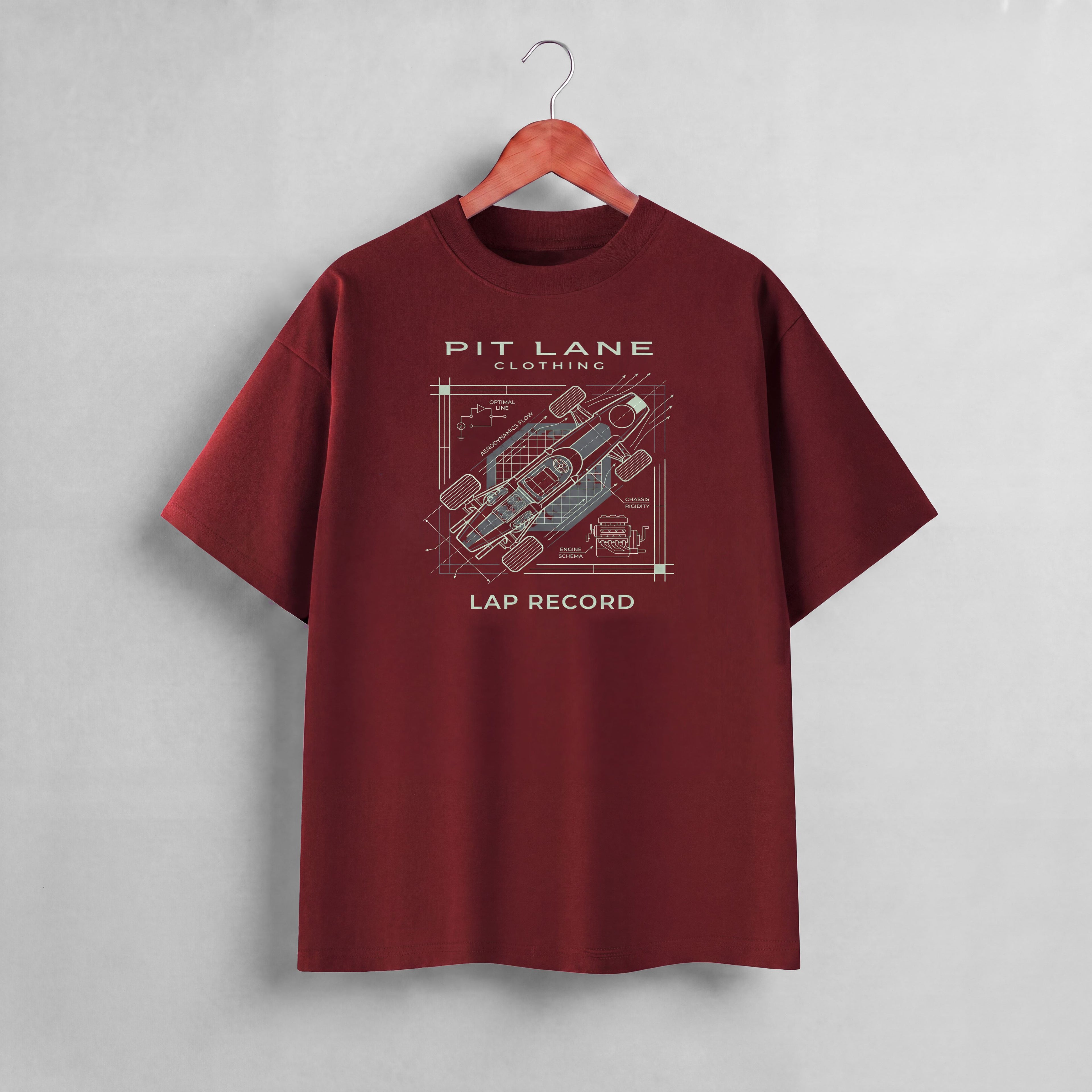 A maroon short-sleeve t-shirt hanging on a wooden hanger against a textured gray wall. The shirt features a graphic print on the front with the text 'PIT LANE CLOTHING' and 'LAP RECORD' surrounding a technical schematic of a race car.